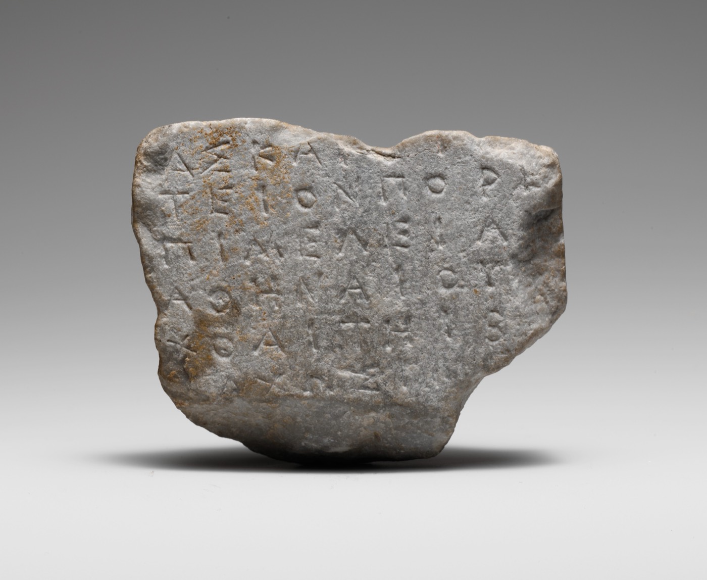 A broken slab of grey marble incised with six lines of Greek capitals. There are no spaces between the words, no lowercase letters and no accent marks; the right side of every line is missing where the stone broke.